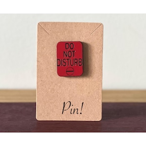Puede incluir: Un pin cuadrado rojo con las palabras "DO NOT DISTURB" en letras negras. El pin está montado en una tarjeta marrón con la palabra "Pin!" impresa en escritura negra. El pin es un accesorio decorativo.