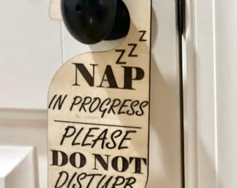 Puede incluir: Un colgador de puerta de madera clara con un recorte circular negro en la parte superior. El texto dice NAP IN PROGRESS PLEASE DO NOT DISTURB con tres Z sobre la palabra NAP. El letrero cuelga de un pomo de puerta negro.