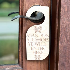 May include: A wooden door hanger with the engraved text "ABANDON ALL SHOES YE WHO ENTER HERE." The sign is light-colored with a floral design and hangs on a dark brown door with a metal handle.
