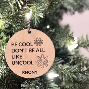 Puede incluir: Un adorno navideño redondo de madera con el texto "Be cool don't be all like... uncool" y el nombre "Rhony" en la parte inferior. El adorno está decorado con diseños de copos de nieve.