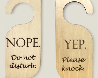 Puede incluir: Dos colgadores de puerta de madera con un gancho en la parte superior. El colgador de la izquierda dice "NOPE. No molestar." El colgador de la derecha dice "YEP. Por favor, llame a la puerta."