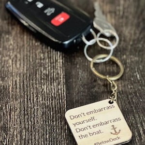 Op de afbeelding: Een zwarte autosleutel met een rode knop en een houten sleutelhanger. De sleutelhanger heeft de tekst "Don't embarrass yourself. Don't embarrass the boat." met een anker en de hashtag #BelowDeck.