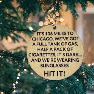 May include: A round, wooden Christmas ornament with the text "IT'S 106 MILES TO CHICAGO...AND WE'RE WEARING SUNGLASSES HIT IT!" in brown lettering. The ornament hangs from a red and white string, set against a blurred Christmas tree background.