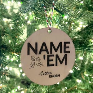 Puede incluir: Un adorno navideño redondo de madera con el texto "NAME 'EM -Sutton RHBOH" en letras negras. Una pequeña rama de acebo está dibujada debajo del texto.