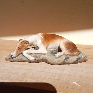 May include: A ceramic figurine of a greyhound dog, lying down on a stone-like base. The dog has a white body with brown markings on its head and back. The figurine is detailed and appears to be handcrafted.