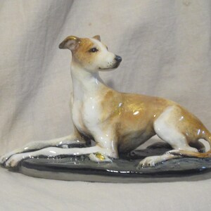 May include: A ceramic figurine of a greyhound dog in a reclining pose. The dog is primarily tan and white, with a glossy finish. It rests on a dark gray oval base, showcasing a detailed and elegant design.