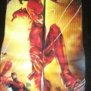 May include: A pair of socks with a colorful illustration of Daredevil, a superhero known for his red suit and heightened senses. The socks feature multiple images of Daredevil in different poses, including a close-up of his face and a full-body shot of him in action.