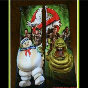 May include: A pair of black and green socks with a Ghostbusters theme. The socks feature images of the Ghostbusters characters, the Stay Puft Marshmallow Man, and Slimer. The socks also have the Ghostbusters logo and the text "Ghostbusters" on them.