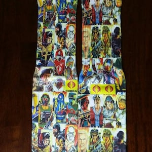 May include: A pair of white socks with a colorful collage of cartoon characters. The socks feature a variety of characters, including a snake, a man with a beard, and a man with a mustache. The socks are printed with the words "Cobra" and "Cobra Commander".