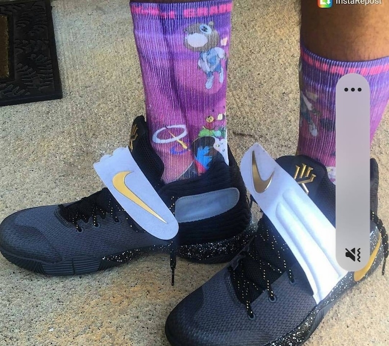 Pode incluir: Um par de sapatos de basquete preto com detalhes brancos e um logotipo Swoosh dourado. Os sapatos t&ecirc;m uma parte superior de malha preta e uma entressola salpicada. Os sapatos s&atilde;o usados com um par de meias roxas e brancas com um personagem de desenho animado.