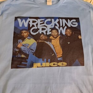 May include: A light blue t-shirt with a graphic print of the rap group "Wrecking Crew" and the word "Juice" in yellow letters. The image features four men in black and blue clothing.