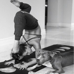 May include: A woman in black athletic wear performs a yoga pose on a black yoga mat with a moon phase design. A gray and white dog lies on the mat next to her.