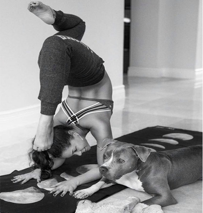 May include: A woman in black athletic wear performs a yoga pose on a black yoga mat with a moon phase design. A gray and white dog lies on the mat next to her.