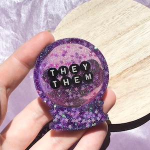 Puede incluir: Presiona papeles de resina con purpurina morada con una cúpula transparente y cuentas de letras negras que deletrean "THEY THEM".