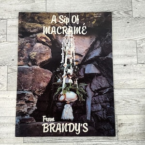 May include: A vintage magazine cover titled "A Sip of Macramé" from Brandy's. The cover features a macramé hanging planter with a white pot, greenery, and a decorative tassel. The background is a stone wall.