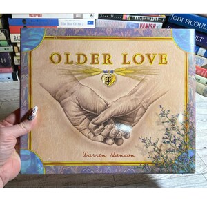 May include: A book cover with the title "OLDER LOVE" in gold lettering. The cover features an illustration of two hands clasped together, with a wheat and heart design above. The author's name, Warren Hanson, is below the illustration. The book is surrounded by other books.