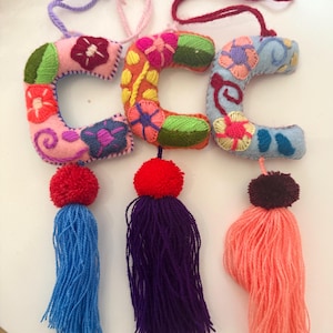 May include: Handmade felt letter ornaments in pink, orange, and blue, each embroidered with colorful floral designs. Each letter has a tassel in blue, purple, and coral. The ornaments have a loop for hanging.