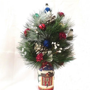 Puede incluir: Un centro de mesa de árbol de Navidad con ramas de pino, campanas rojas y azules, muñecos de nieve blancos, piñas y flores rojas en un jarrón de cerámica blanco con una escena de invierno.