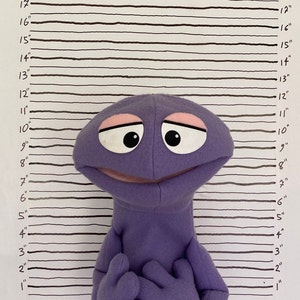May include: A purple puppet with a big smile and large eyes. The puppet is standing in front of a white background with black lines.