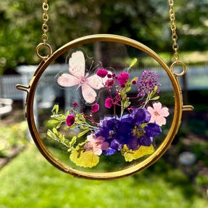 May include: A round, gold-colored pendant with a clear glass center, showcasing a delicate arrangement of dried flowers and a butterfly. The flowers are in shades of purple, pink, and yellow, with a gold chain for hanging.