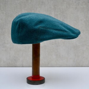Puede incluir: Una gorra plana de lana azul verdosa con una costura en la corona. La gorra se muestra en un soporte de madera con una base roja.