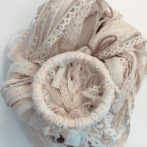 Puede incluir: Un montón de encaje blanco y crema, cinta y recortes de tela atados con cuerda. El encaje está dispuesto en forma circular con un pequeño anillo de madera en el centro.