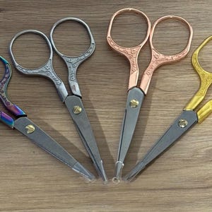 Könnte beinhalten: Vier Paare dekorativer Stickschere in verschiedenen Farben. Die Scheren haben verzierte Griffe in schillernd, silber, kupfer und gold. Die Klingen sind silberfarben und die Scheren sind auf einer Holzoberfläche angeordnet.