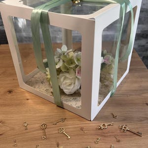 May include: A square white gift box with clear windows on the sides and top, tied with a sage green ribbon. Inside, a bouquet of white and pale green artificial flowers rests on a floral-patterned base. Gold-toned skeleton keys are scattered.