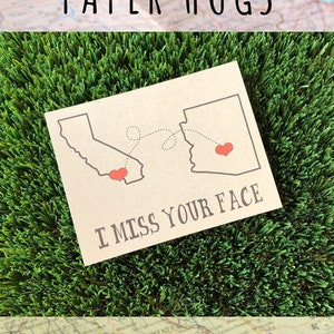 May include: A rectangular tan greeting card featuring a dark gray outline of California and Arizona. A dashed line connects the two states, each marked with a small red heart. The text I MISS YOUR FACE is printed in dark gray below the maps.