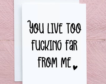 Può includere: Un biglietto d'auguri bianco con il testo nero "YOU LIVE TOO FUCKING FAR FROM ME" seguito da un piccolo cuore nero. Il biglietto è posizionato davanti a una busta bianca semplice, presentando un design pulito, essenziale e ben leggibile.