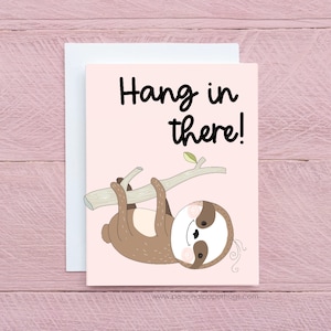 May include: A greeting card with a light pink background and a cartoon sloth hanging from a branch. The text "Hang in there!" is written in black script above the sloth. A white envelope is behind the card.