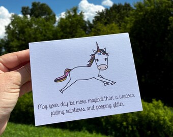 May include: A white greeting card featuring a cartoon unicorn with a colorful mane and tail. The text printed below the unicorn reads: May your day be more magical than a unicorn farting rainbows and pooping glitter. The card has a smooth finish.