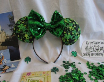Peut inclure: Oreilles de souris Minnie vertes et noires avec un nœud en sequins verts. Les oreilles sont recouvertes d'un imprimé trèfle. Le serre-tête est noir.