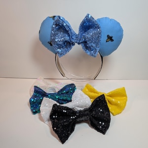 May include: A set of five sequined bows in various colors, including blue, teal, white, black, and yellow. The bows are arranged in a semi-circle on a white surface.