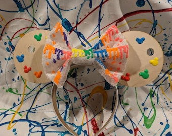 May include: A pair of white mouse ears with a colorful paint splattered bow. The ears are shaped like paint palettes with Mickey Mouse silhouettes painted on them.