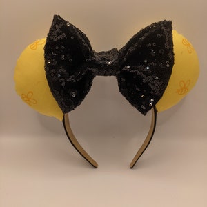 May include: Yellow fabric mouse ears with a black sequin bow. The ears have a pattern of small yellow bees.