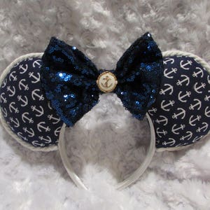 May include: A pair of white mouse ears with a blue and white anchor print fabric. The ears have a large blue sequin bow with a gold anchor button in the center.