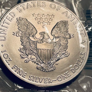 K&ouml;nnte beinhalten: Eine silberne American Eagle-M&uuml;nze mit der Aufschrift "United States of America 1 oz. Fine Silver - One Dollar" und einem Wei&szlig;kopfseeadler mit ausgebreiteten Fl&uuml;geln, der ein Schild mit dreizehn Streifen und einen Olivenzweig in seinen Krallen h&auml;lt.