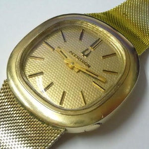 May include: A gold-toned Accutron watch with a square face and a mesh band. The watch face is gold with black markers and hands. The band and watch case are a matching gold color.