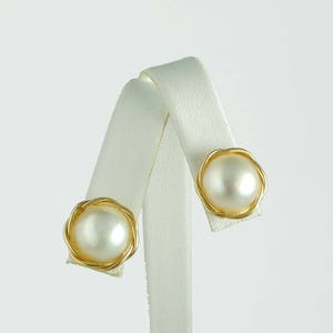 以下が含まれることがあります： 金色のイヤリングのペアで、それぞれに白い真珠がセットされています。真珠はねじれたワイヤーのデザインにセットされています。