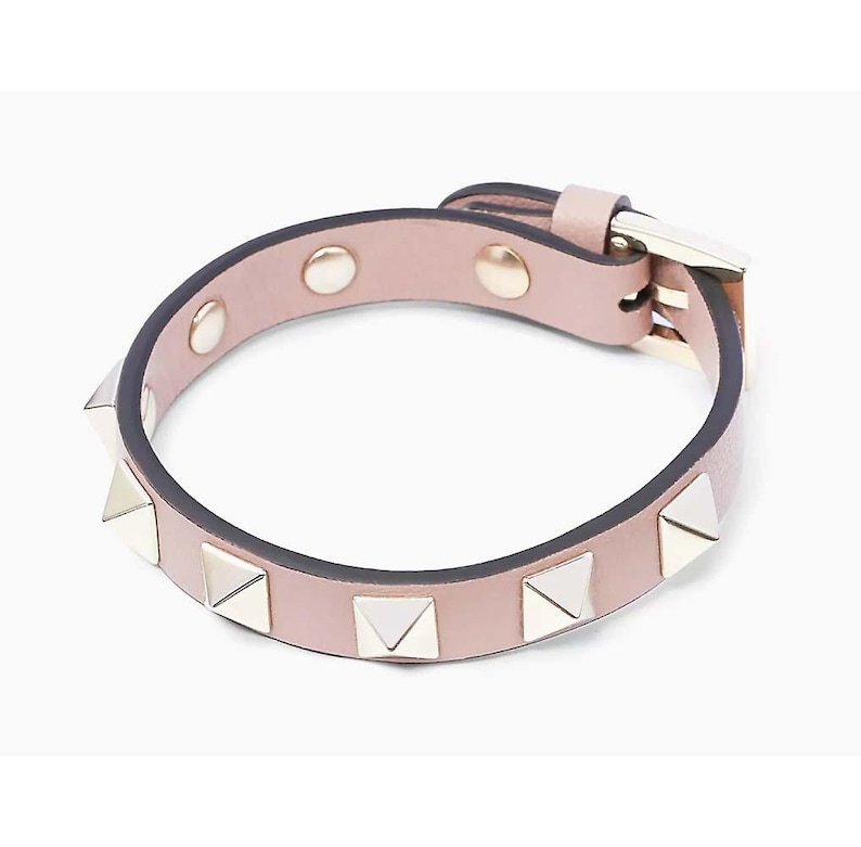 Puede incluir: Un brazalete de cuero rosa claro con tachuelas de pir&aacute;mide plateadas. El brazalete tiene un cierre de hebilla.