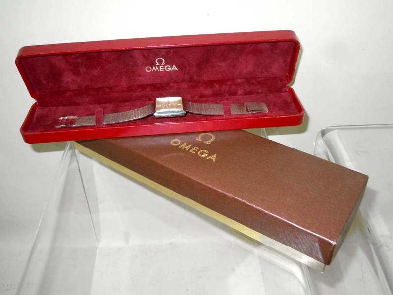 May include: A red and brown watch box with the Omega logo on the lid. The watch is inside the box and has a silver band.