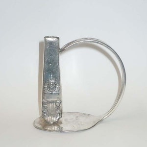 Puede incluir: Una escultura de una cuchara de plata con forma de la letra D. La cuchara se ha doblado y retorcido para crear la forma. El mango de la cuchara es la parte vertical de la D, y el cuenco de la cuchara es la parte curva de la D.