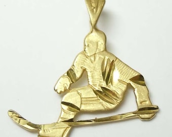 Könnte beinhalten: Ein goldfarbener Metallanhänger in Form eines Eishockeyspielers in Laufposition, der einen Eishockeyschläger hält. Die Oberfläche weist texturierte Diamantschliff-Details auf Trikot und Hose auf, mit einer dreieckigen Öse zum Aufhängen.
