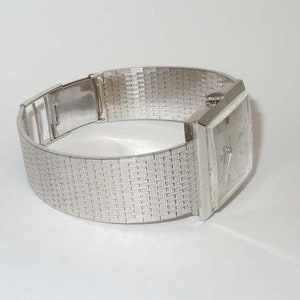 May include: A silver square-shaped wristwatch with a silver metal band. The band has a woven pattern.