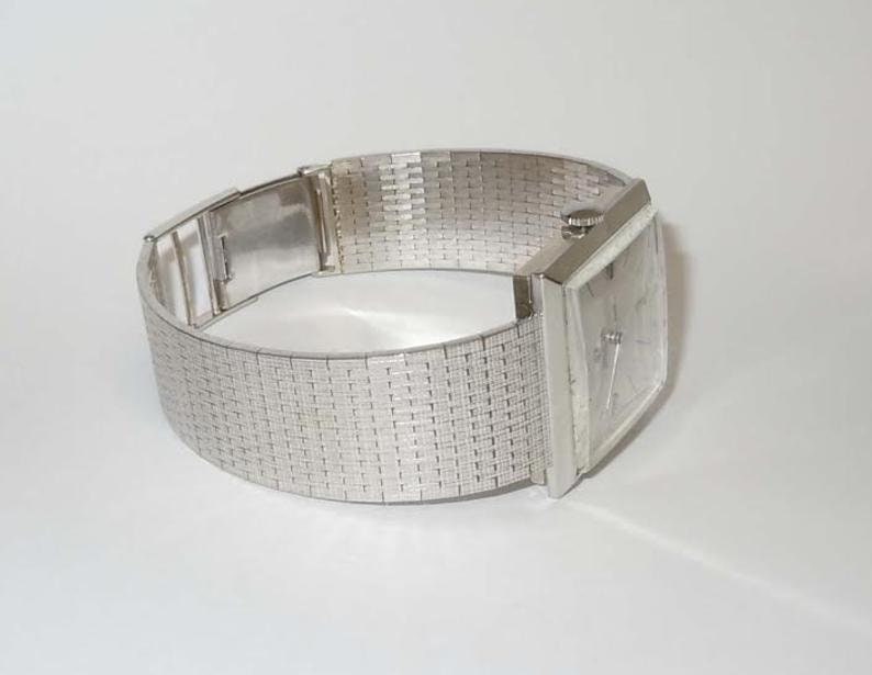 May include: A silver square-shaped wristwatch with a silver metal band. The band has a woven pattern.