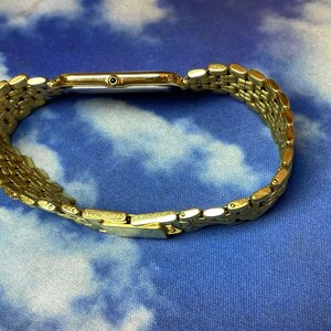 May include: A gold-toned wristwatch with a rectangular face and a linked bracelet. The watch is displayed on a blue and white patterned surface. The watch band is made of small, connected rectangular links.