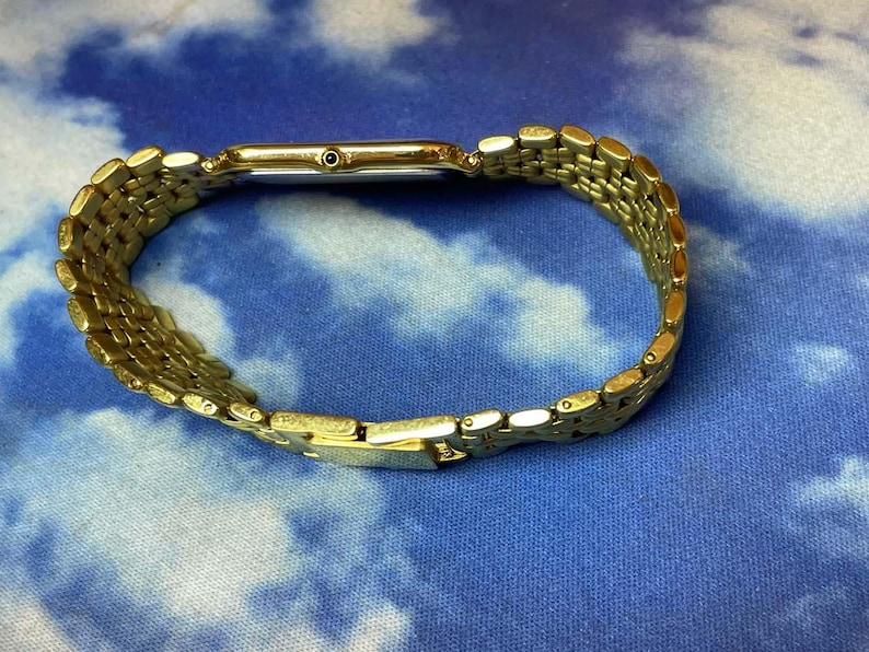 May include: A gold-toned wristwatch with a rectangular face and a linked bracelet. The watch is displayed on a blue and white patterned surface. The watch band is made of small, connected rectangular links.