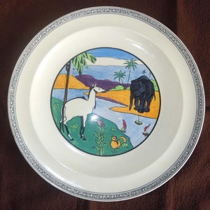 May include: A cream-colored decorative plate with a detailed border. The center features a colorful illustration of a white animal, an elephant, palm trees, a river, and small boats. The plate has a vintage aesthetic.