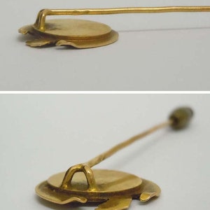 May include: A gold-toned pin with a round, slightly raised, circular design. The pin has a long, thin, straight shaft.